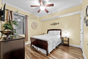 Un dormitorio con cama de madera, ventilador de techo, mesita de noche con lámpara, un adorno en la pared y una ventana con persianas.