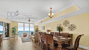 A spacious dining area with a large wooden table and chairs, a ceiling fan, and a view of the ocean through large windows. - Sienna on the Coast 1006 | Enjoy Beautiful 10th Floor Views of the Gulf! (Gulfport)