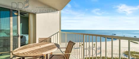 A balcony with a table and chairs, overlooking a beach and ocean.