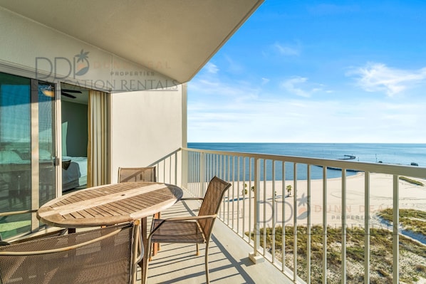 A balcony with a table and chairs, overlooking a beach and ocean.