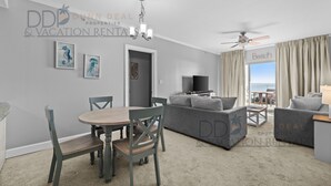 A modern living room with a dining area, grey sofas, a flat-screen TV, and a balcony with a view. - Sea Breeze 907 | Condo Directly on the Beach! (Biloxi)