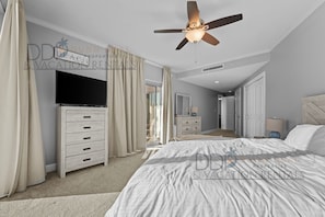 A bedroom with a bed, a ceiling fan, a TV, a dresser, and a door leading to another room.
