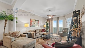A spacious living room with a ceiling fan, a large window, and a view of the outdoors. There are multiple seating areas, including a sofa, a chair, and a red ottoman. - Legacy Tower I 1403 | Penthouse Unit Overlooking the Beautiful Gulf! (Gulfport)