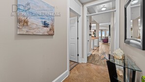 A hallway with a beach-themed painting, a glass table with decorative items, and a view into a living area with a purple chair. - Legacy Tower I 1403 | Penthouse Unit Overlooking the Beautiful Gulf! (Gulfport)
