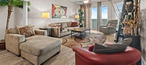 A spacious living room with a ceiling fan, a large window, and a view of the outdoors. There are multiple seating areas, including a sofa, a chair, and a red ottoman.