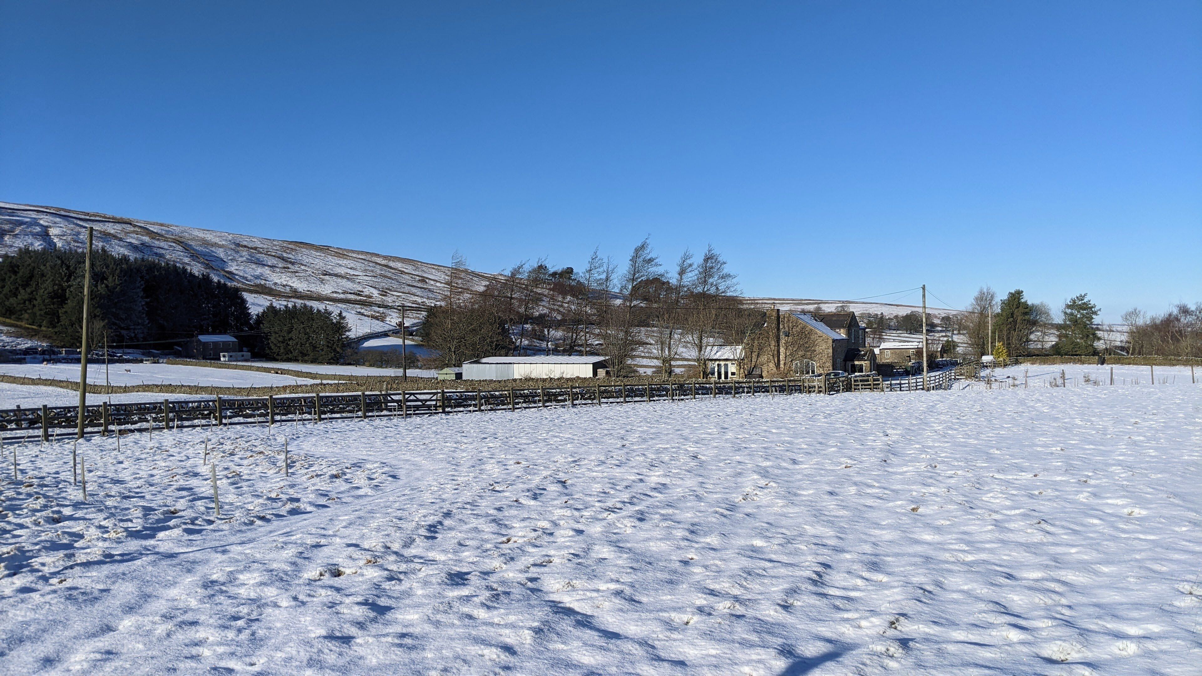 Paisatge rural amb un camp cobert de neu, una tanca de fusta i alguns edificis al fons.
