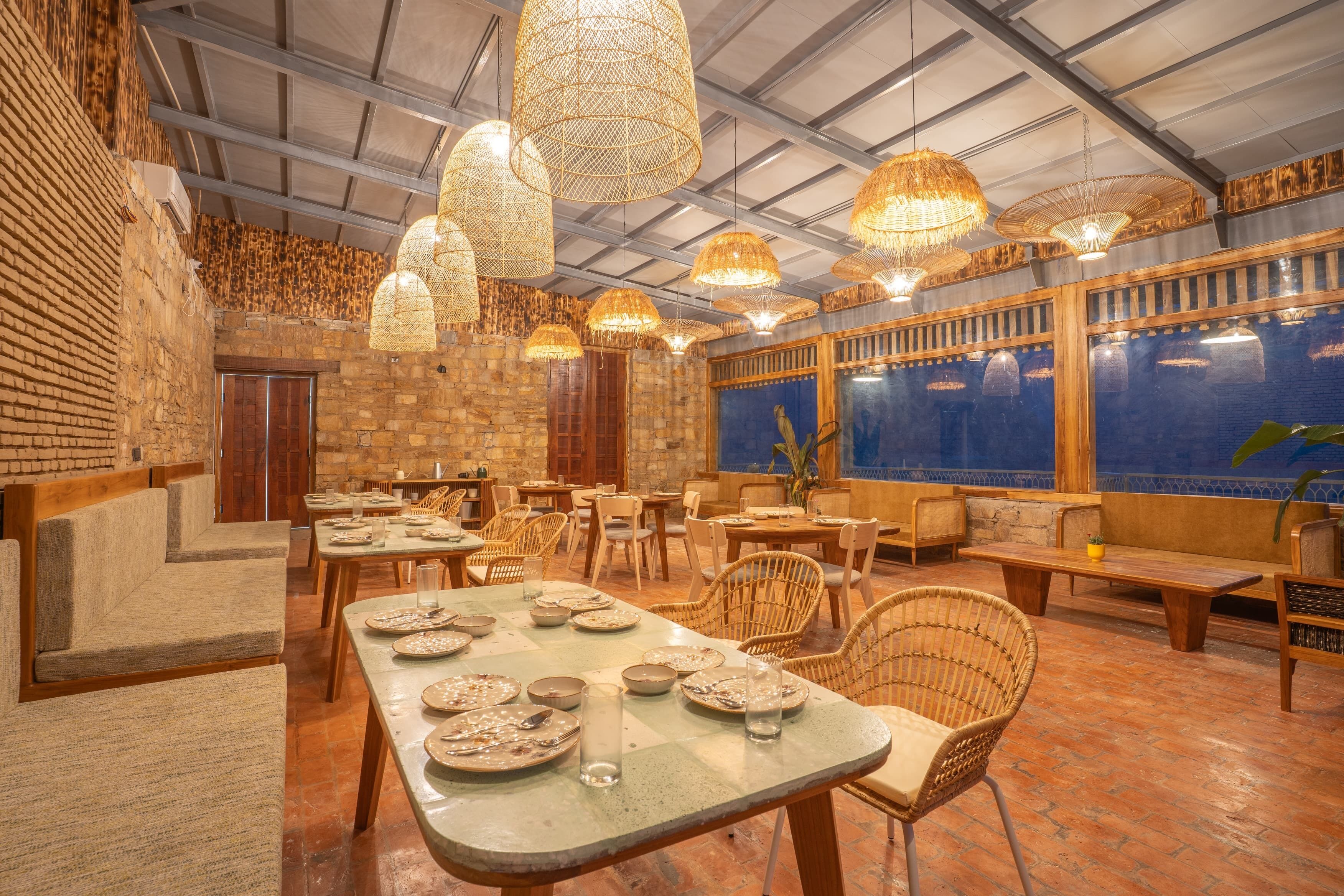 Un restaurante con paredes de ladrillo, muebles de madera y lámparas de mimbre colgantes. Las mesas están preparadas con platos y vasos.