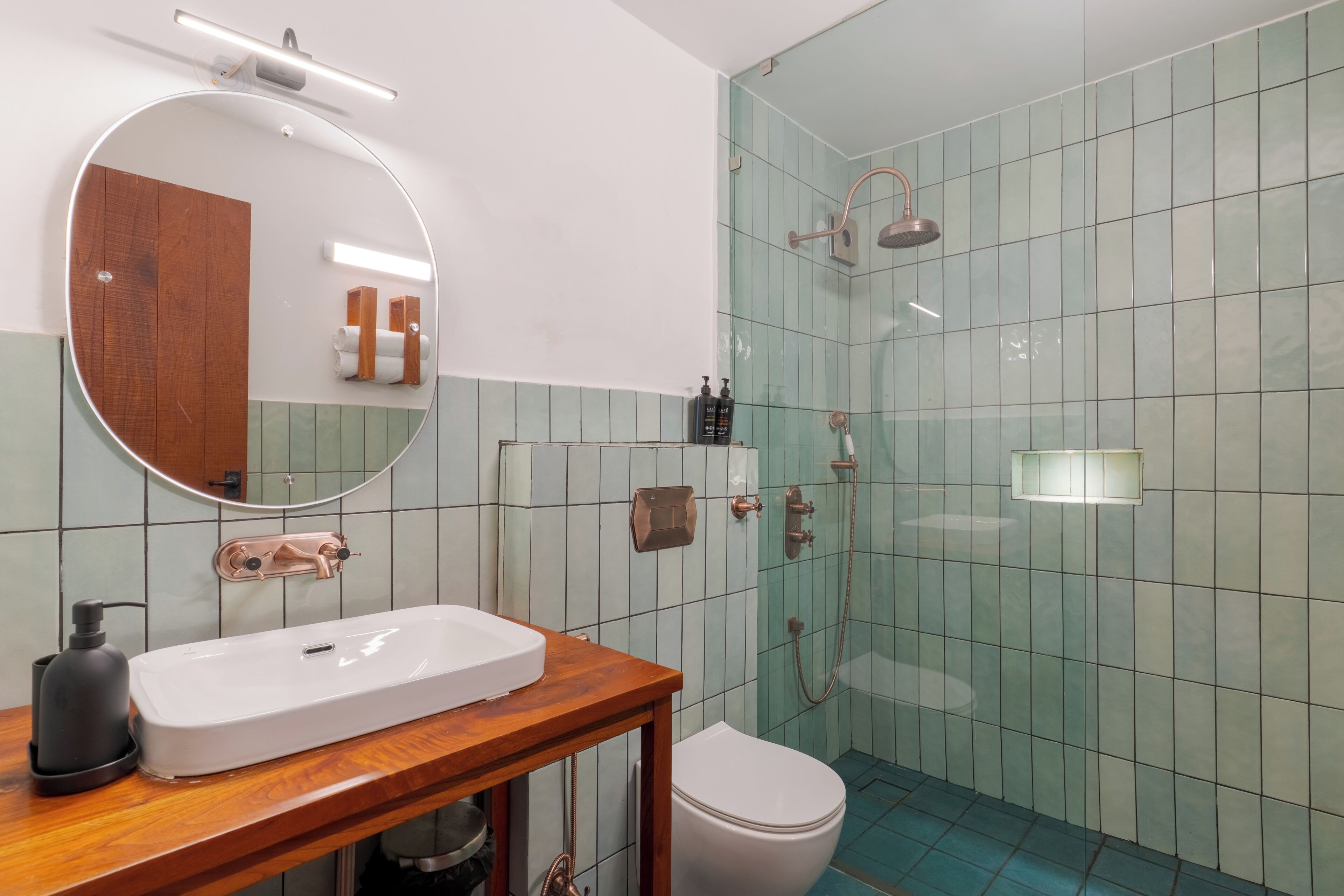 A bathroom with a wooden vanity, a round mirror, a white sink, a toilet, and a glass shower enclosure.