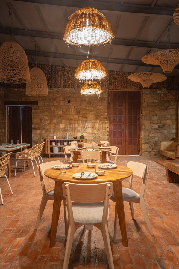 A dining area with round wooden tables, wicker hanging lamps, and stone walls.