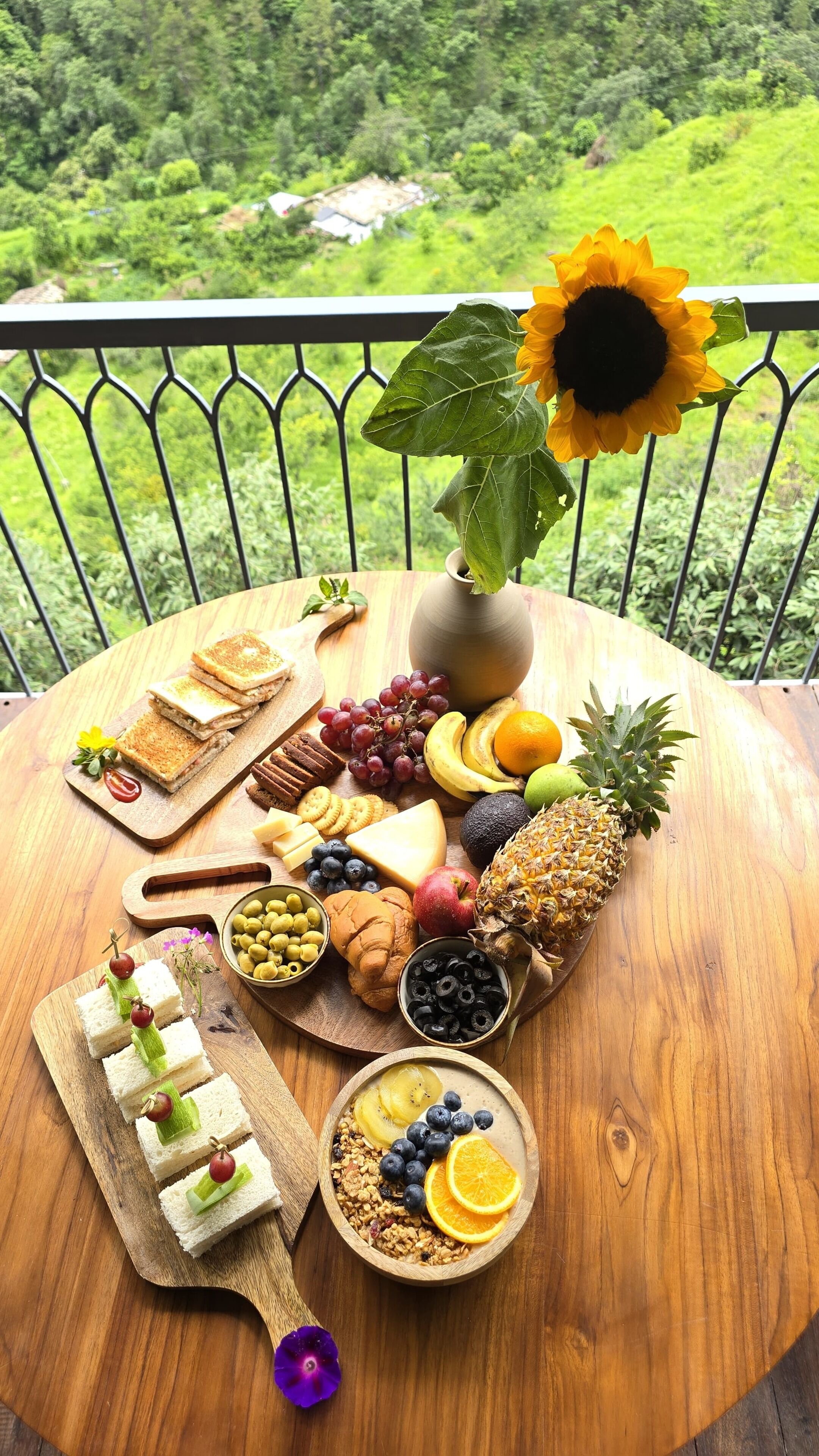A wooden table with a variety of food items and a sunflower, set against a balcony railing with a view of greenery.