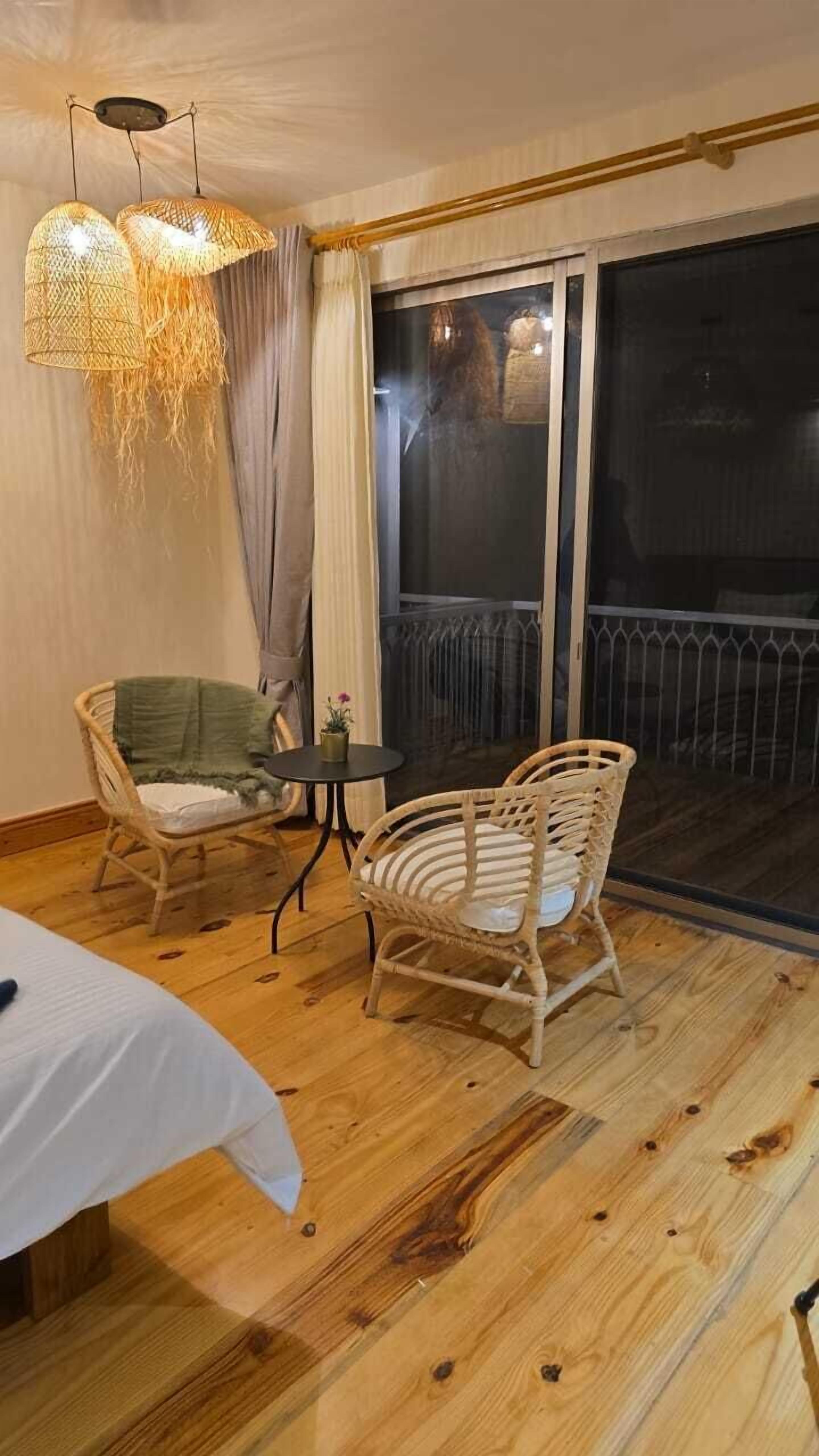 A room with a wooden floor, two wicker chairs, a small round table, and a bed with a white bedspread.
