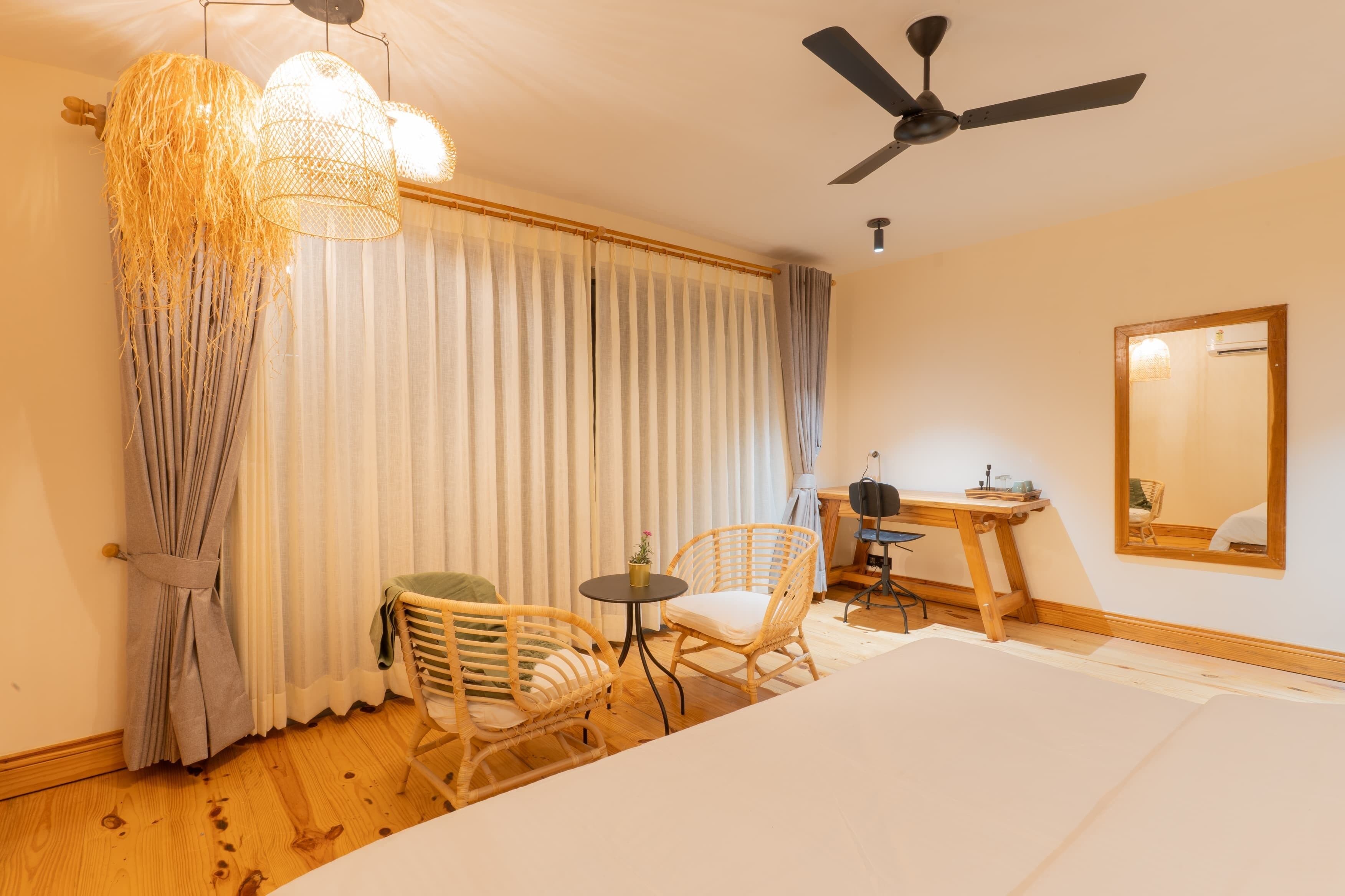 A hotel room with a bed, two wicker chairs, a small table, a desk, a ceiling fan, and a large window with curtains.