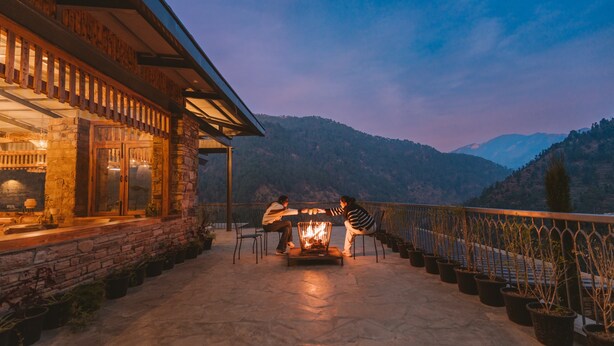 A stone building with a fire pit and chairs on a patio overlooking mountains. - Over The World (Kosiyakutoli)