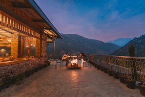 A stone building with a fire pit and chairs on a patio overlooking mountains. - Over The World (Kosiyakutoli)