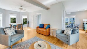 Ein geräumiges Wohnzimmer mit hoher Decke, Holzboden und einem großen Fenster, das für natürliches Licht sorgt. Man sieht zwei Sofas, einen Couchtisch und einen Essbereich.