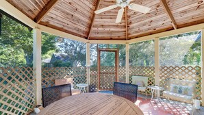 Ein überdachter Außenbereich mit Holztisch und Stühlen, einem Deckenventilator und einem Gitterzaun.