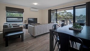 A modern living room with a sofa, a TV, a dining table with chairs, and a balcony with a view of trees. - Long Island Retreat: Pelican. The Perfect Riverside Family Getaway. (Murray Bridge)