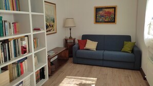 Een woonkamer met een blauwe bank, een witte boekenkast, een schilderij en een lamp.
