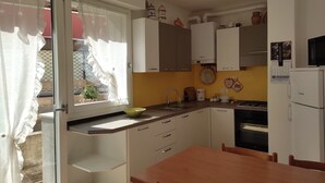 Een kleine keuken met een spoelbak, fornuis en kastjes, een eettafel en een raam met gordijnen.