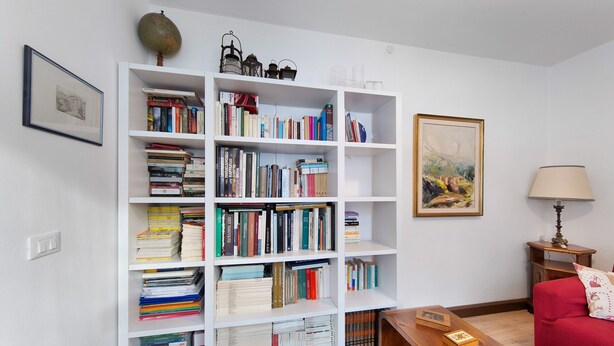 Een kamer met een witte boekenkast vol boeken, een rode bank, een houten bijzettafeltje en een lamp.