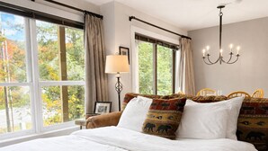 Ein großes Bett mit weißer Bettwäsche, eine Kristallleuchter, eine Lampe und Blick auf Bäume durch große Fenster.