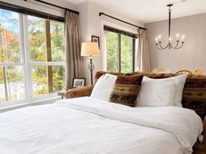 Een groot bed met witte lakens, een kroonluchter, een lamp en uitzicht op bomen door grote ramen.