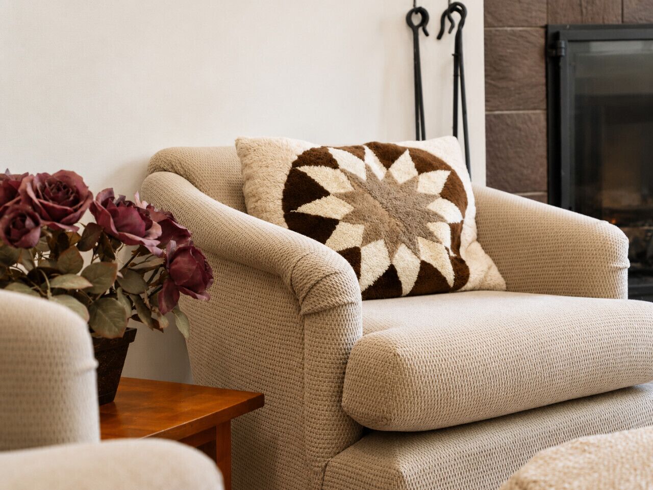A beige armchair with a patterned throw pillow, a wooden side table, and a vase with flowers.