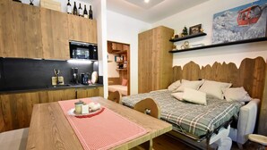 Une pièce compacte comprenant une table en bois, un lit avec une couette à carreaux, une armoire intégrée, une petite kitchenette et une étagère avec des livres et un tableau.