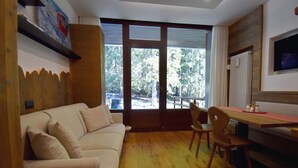 Un salon chaleureux avec un canapé beige, un plancher en bois, une table à manger avec des chaises, un téléviseur fixé au mur et de grandes fenêtres donnant sur des arbres et une voiture à l’extérieur.