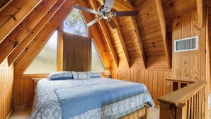 Ein Schlafzimmer mit geneigter Decke, Holzwänden, einem Bett mit gemusterten Textilien, einem Deckenventilator und einem Fenster mit Blick auf Bäume.