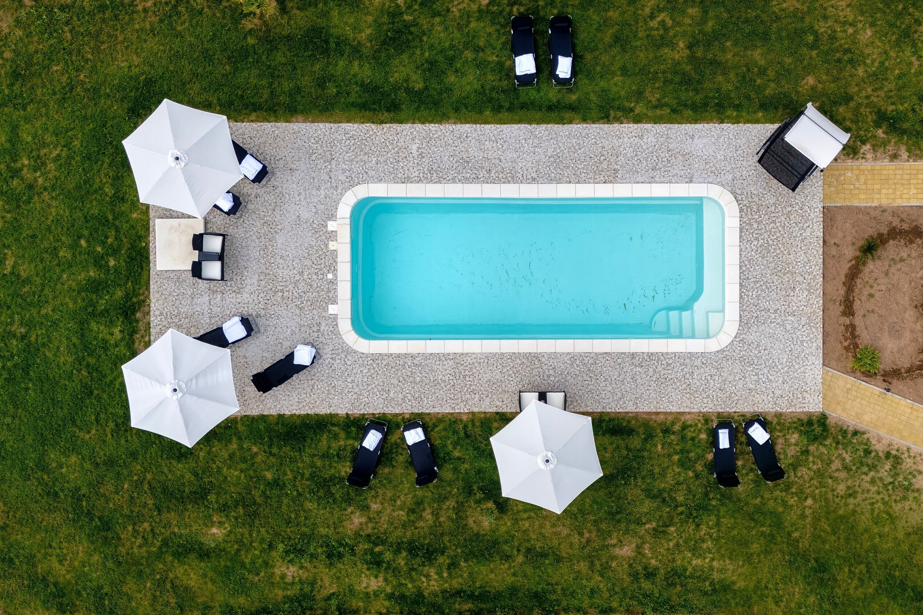 Ein Luftbild eines rechteckigen Schwimmbeckens, umgeben von Liegestühlen und Sonnenschirmen, mit einem gepflasterten Weg und Rasenflächen.