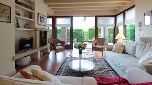 Ein geräumiges Wohnzimmer mit einem weißen Sofa, einem Couchtisch und Korbstühlen. Ein Fernseher steht auf einem Regal, und ein großes Fenster bietet Blick ins Grüne.