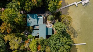 An aerial view of a house surrounded by dense trees, with a dock extending into a body of water. - Lakeside Escape | 5BR | Hot Tub | Family Friendly (Traverse City)
