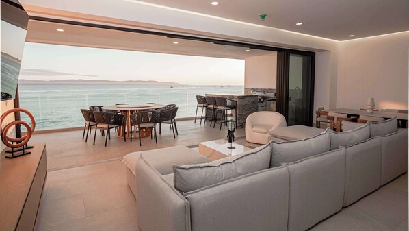 Ein modernes Wohnzimmer mit einem großen grauen Sofa, einem runden Esstisch mit Stühlen und Meerblick.