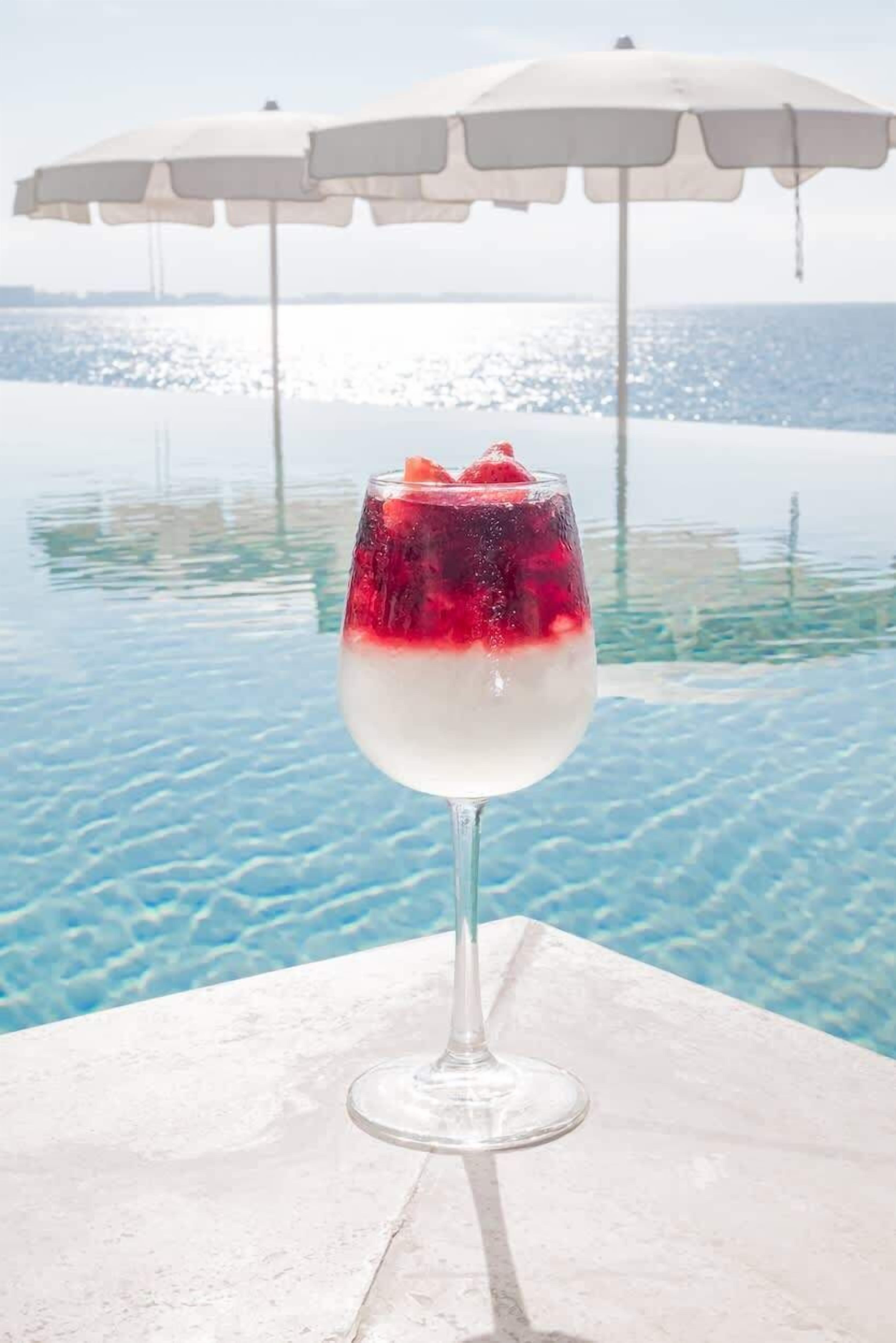 Un vaso de cóctel con una bebida en capas y una fresa como decoración, sobre una mesa junto a una piscina con una sombrilla blanca al fondo.