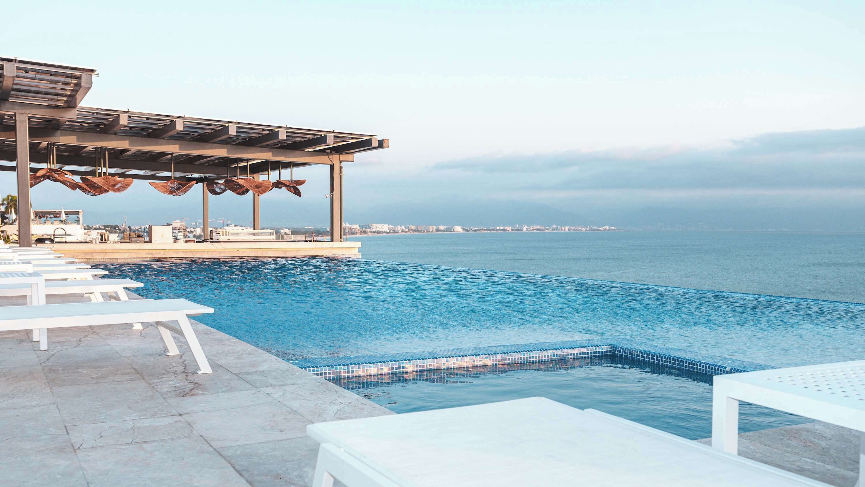 A pool area with white lounge chairs, a covered seating area with umbrellas, and a view of the sea.