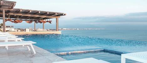 Zona de piscina con sillones blancos, un área de estar cubierta con sombrillas y vista al mar.