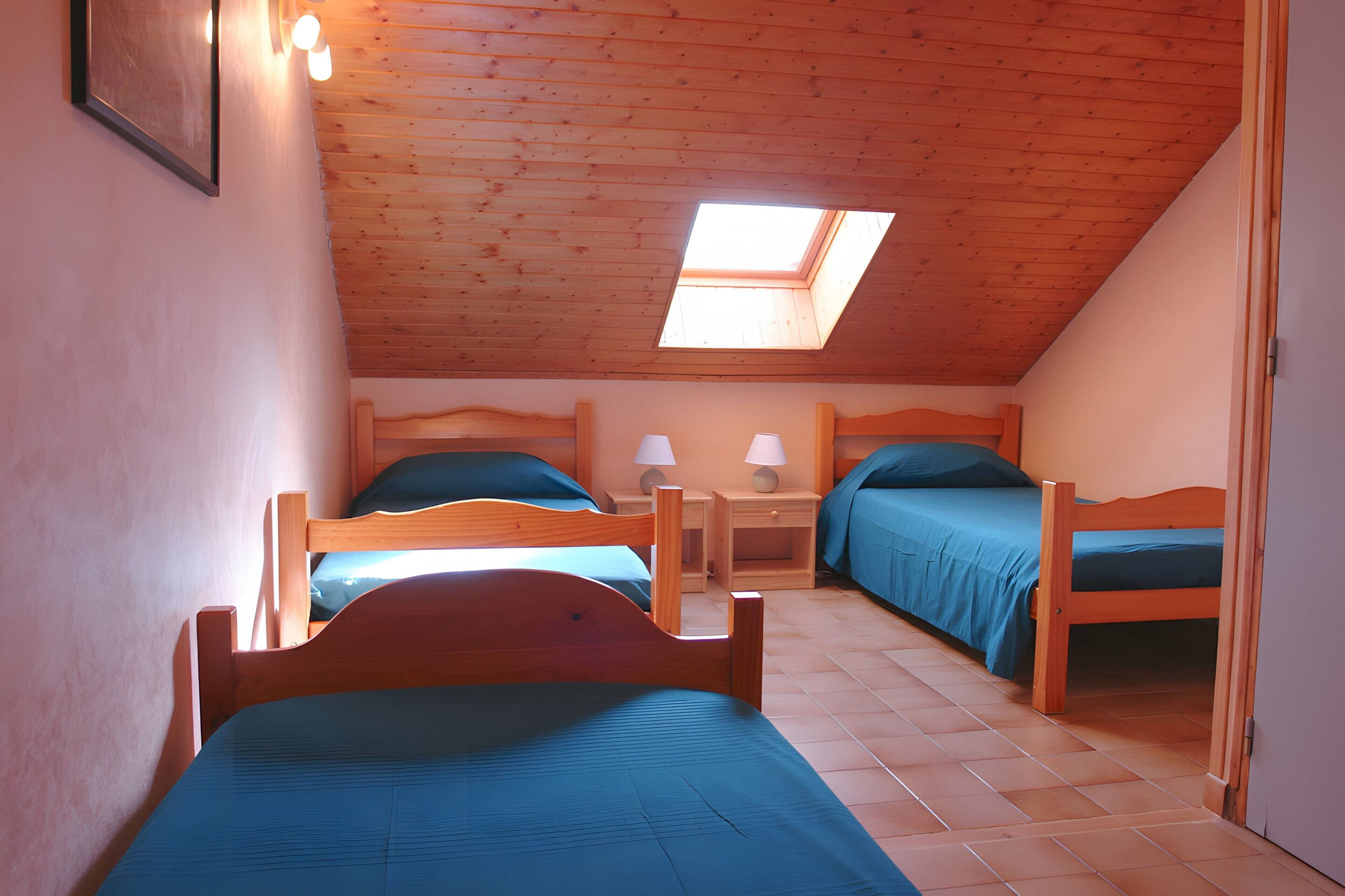 Quarto com duas camas de madeira, um claraboia e piso ladrilhado.