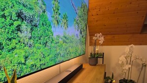 Un interno moderno con una grande TV a schermo piatto che mostra un paesaggio tropicale, soffitto in legno e una reception in legno con candele, piante in vaso e un blocco note.