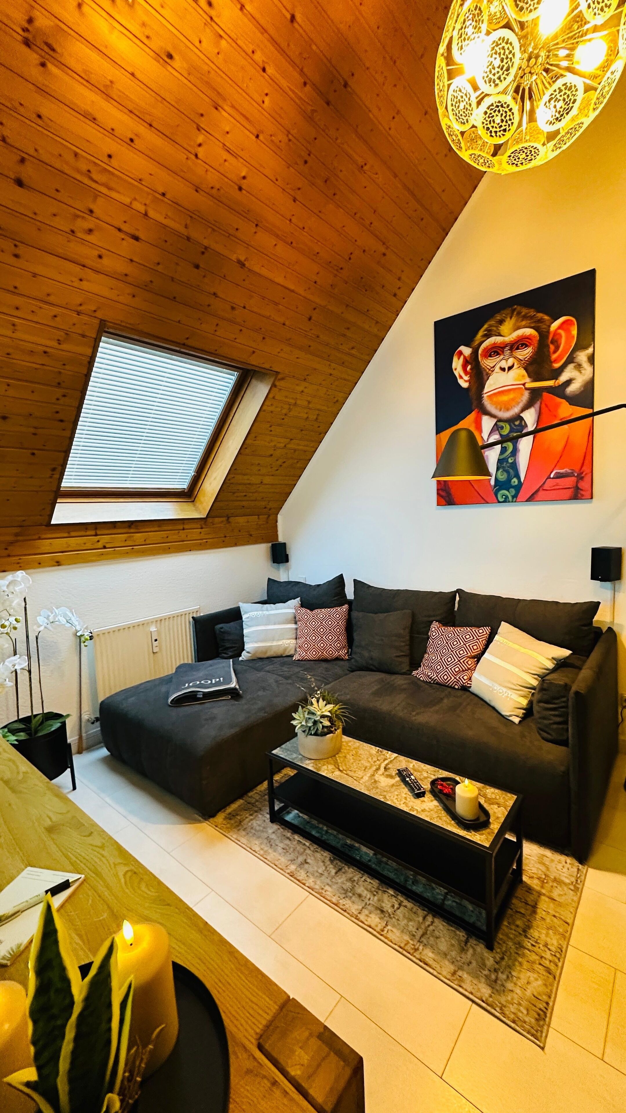 Un salon chaleureux de style industriel, avec un plafond en pente, une grande fenêtre, un ensemble de meubles modernes comprenant un canapé, une table basse et un tableau accroché au mur.