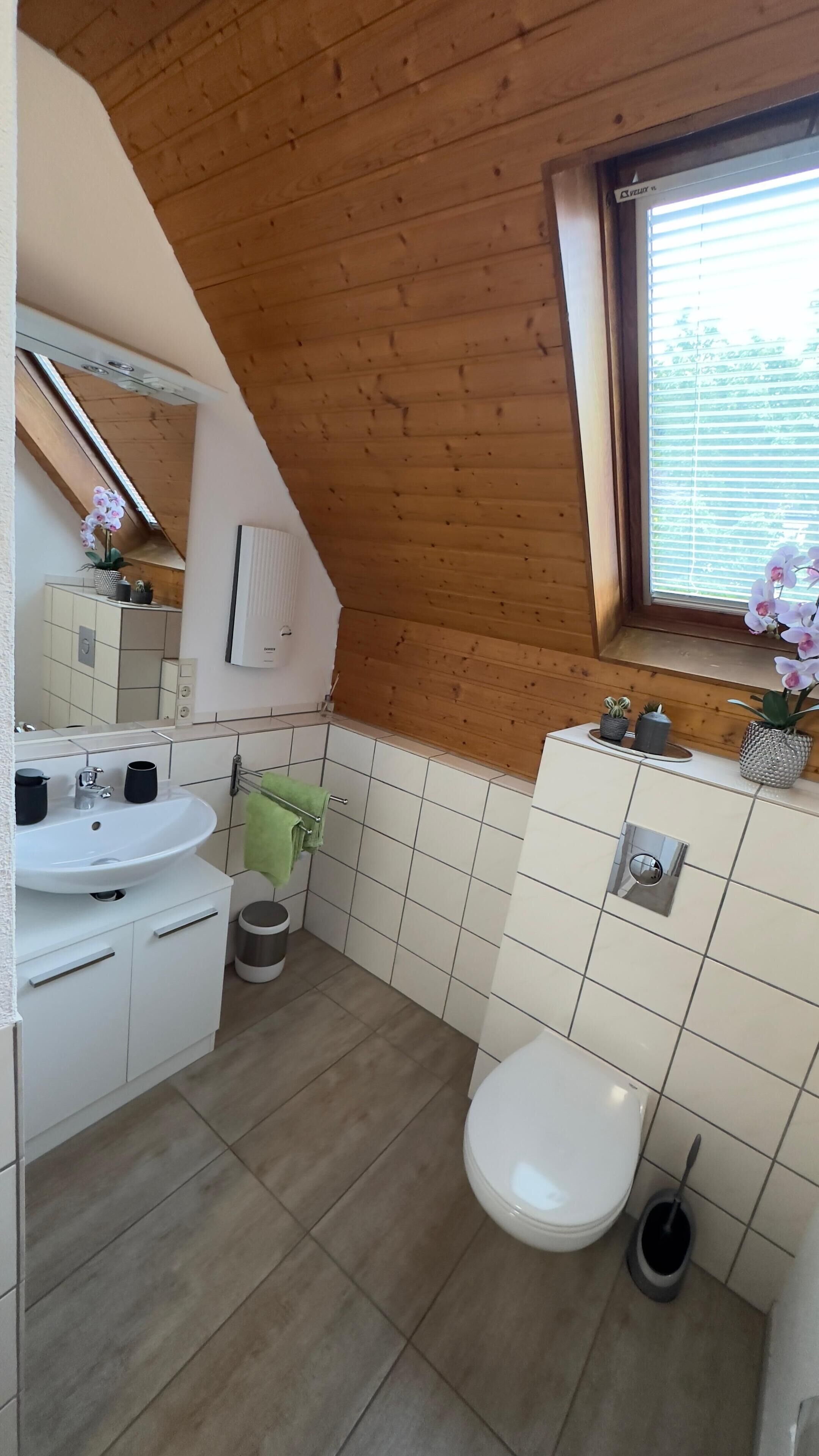 A small, compact bathroom with a wooden ceiling, white tiles, a sink, a toilet, and a window with blinds.