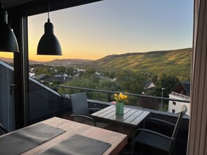 Un balcón con mesa y sillas, con vistas a un paisaje pintoresco de un valle con casas y vegetación.