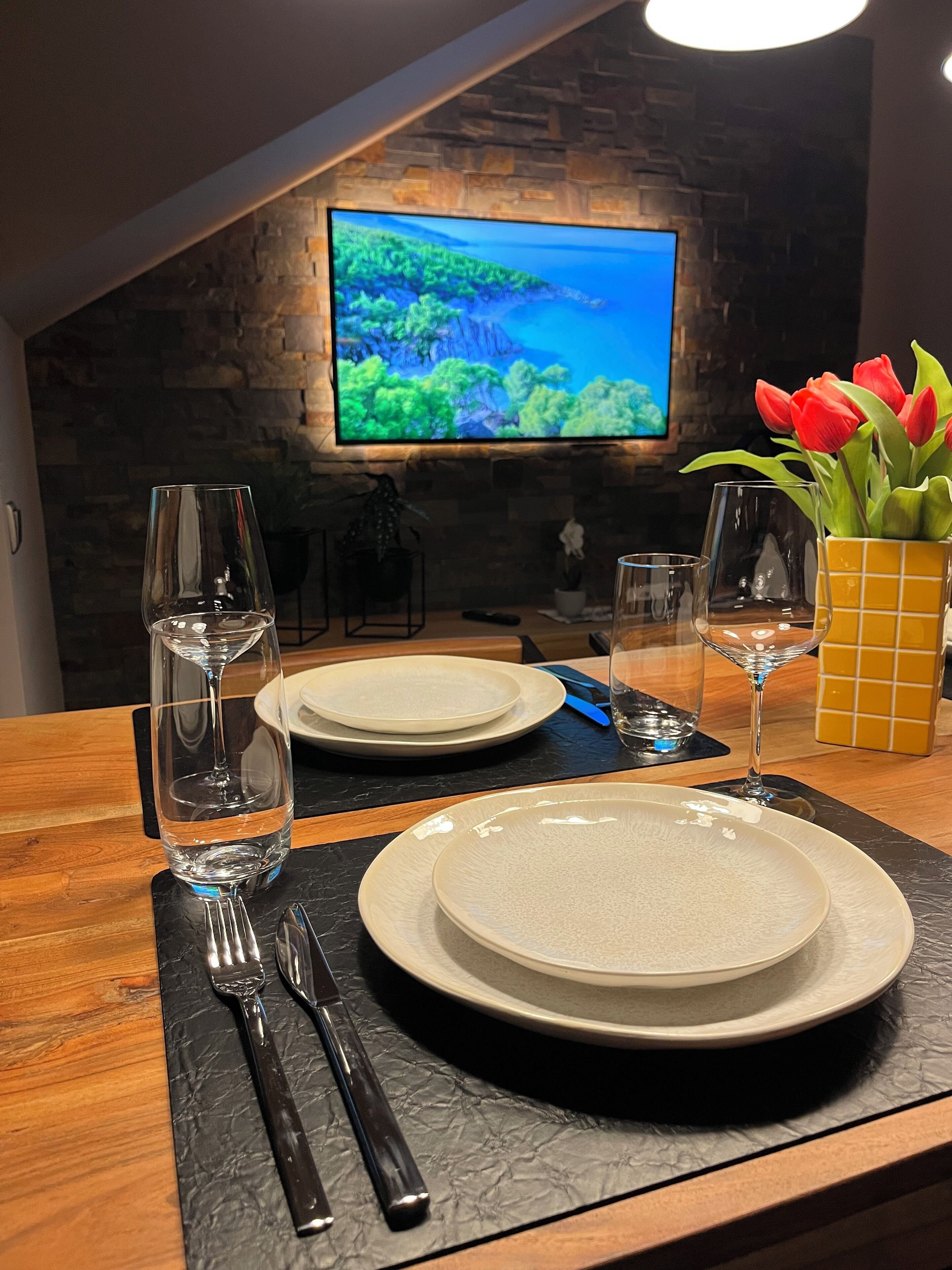 A dining table set with plates, cutlery, and glasses, with a TV displaying a scenic landscape in the background.