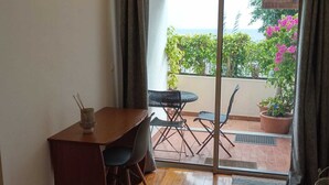A room with a wooden floor, a desk with a chair, a small table with a chair, and a sliding glass door leading to an outdoor area with potted plants and a view of the sea.