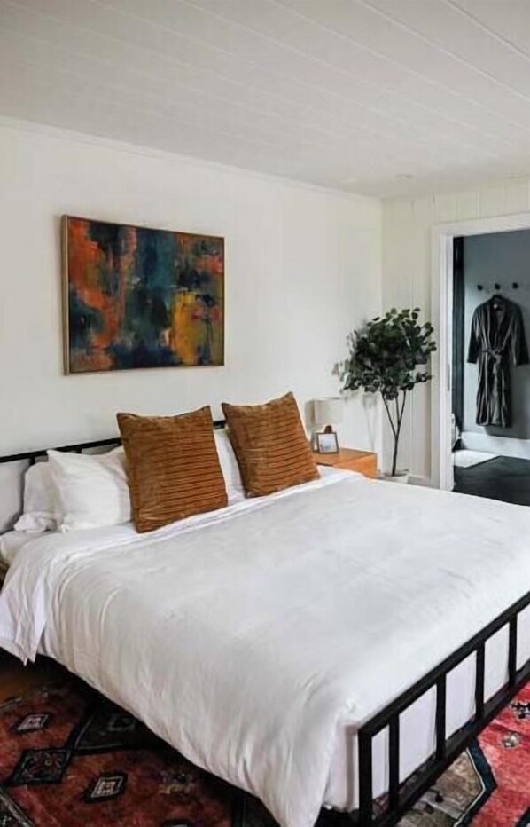 A bedroom with a bed, pillows, a nightstand, a plant, and a door with a robe hanging.