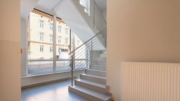 A modern interior with a staircase leading to an upper floor, a large window with a view of a building, and a radiator on the wall. - AS HOME Kościuszki 89 (Wrocław)