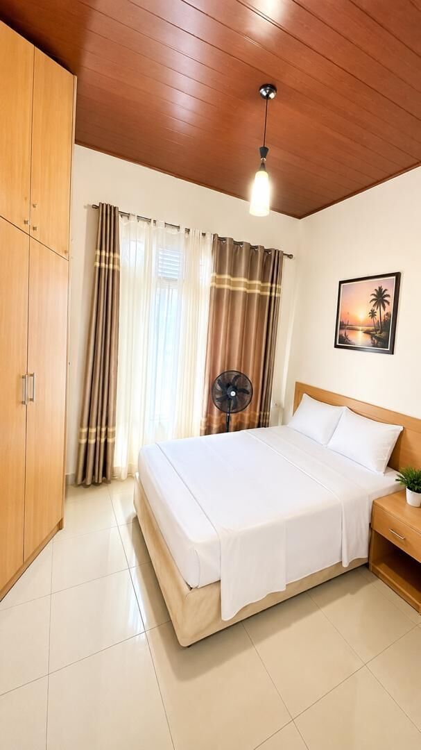 A bedroom with a bed, a nightstand, a fan, and a wooden wardrobe.