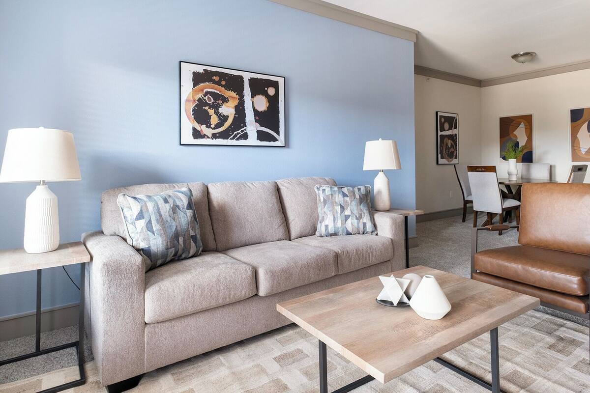 A modern living room with a beige sofa, a wooden coffee table, and a brown leather armchair. There are two floor lamps and abstract art on the walls.