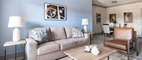 A modern living room with a beige sofa, a wooden coffee table, and a brown leather armchair. There are two floor lamps and abstract art on the walls.