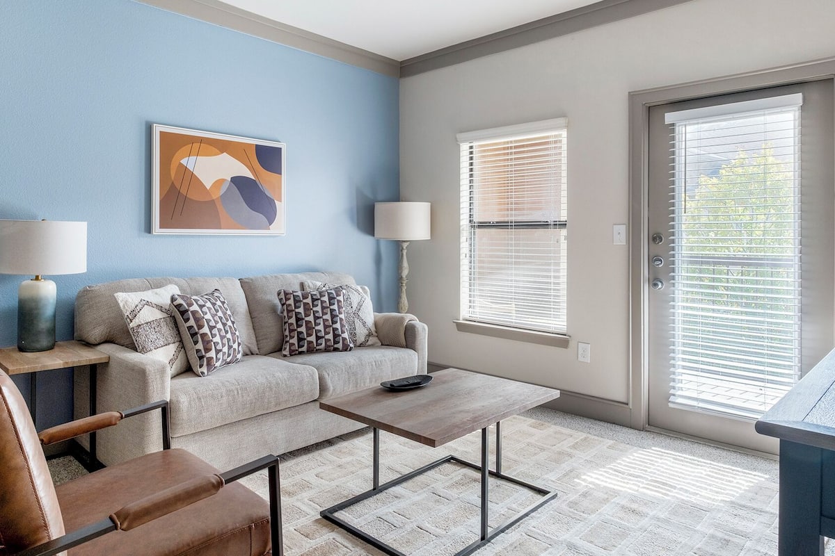 A living room with a sofa, coffee table, and armchair, a painting on the wall, and a door with blinds.